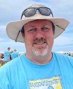 A man smiles for the camera wearing a beige sunhat with sunglasses resting on it and a blue t-shirt with the event poster image printed on it. The beach and spectators are seen in the background.