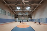 Five people, seen from a distance, play basketball inside a large gym - the windows are barred