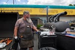 A man wearing a black apron stands between a large grill and a smoker.