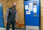 principal wearing a black vest and plaid shirt, walking past a blue classroom door that is marked with a notice stating isolation area