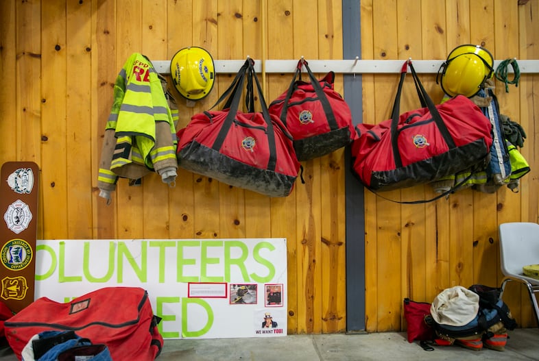 Firefighting equipment hangs above a "Volunteers Needed" sign at Payton Station, headquarters of the Baker Rural Fire Protection District, Baker City, Ore., Aug. 1, 2024. Baker Rural Fire Protection District is a volunteer department that sent crews to the Durkee, Thompson and other local fires throughout July. Chief Sean Lee says, "We're always stretched pretty thin, but this year has been so much more. Our yearly budget starts in July, and we ran out two weeks in." 