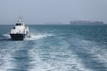 A patrol boat moves through open water as cargo ships sit at anchor behind it.
