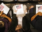 Three people hold U.S. flags as they wait to be sworn in as American citizens at a naturalization ceremony on the Intrepid Sea-Air-Space Museum.