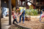 A dad helps his toddler navigate a low playground barrier.  The dad is wearing a tie-dye t-shirt; the little girl holds a red stuffed-animal toy.