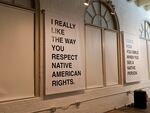 Canvas printed signs in all capitals with messages about respecting Native American people adorn one wall of the exhibition during the “Citizen Fellow: Art as Archive and Memory” exhibition opening at the Center for Native Arts and Cultures on Sept. 19, 2024. The pieces by Anna Tsouhlarakis, citizen of the Navajo Nation with Creek and Greek descent, are part of her series, the Native Guide Project.