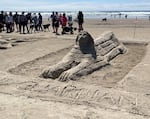 A sand sculpture of a reclining sphinx statue, with spectators in the background.