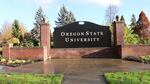 A sign on a brick wall reads "Oregon State University," and is surrounded by trees in the background and a lawn and sidewalk in the foreground.