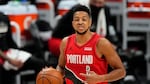 A man in a red and black jersey with the word "Portland" on it has a hand on a basketball on the court.