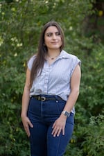 Nusheen Farahani is standing outdoors, with green foliage in the background. She is wearing blue pants and a light-blue sleeveless shirt.
