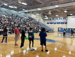 Five students stand on one end of the gymnasium floor at Woodburn High School. One in the center is holding a green balloon. Students in the center of the gymnasium are hugging to pop another balloon. Hundreds of other ninth graders sit in the bleachers in the background.