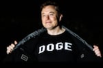 In this photo, Elon Musk is wearing a black shirt with "DOGE" printed on the front in large white capital letters. He's using his hands to hold wide open the dark jacket he's wearing over the shirt. Musk is smiling, and the background behind him is black.