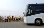 A group of tourists mull in front of a wheat field on the side of a gravel road as the bus driver tries in vain to get the bus moving again from the driver's seat.