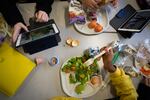 Photographed from above, seventh-graders sit together at a cafeteria table during lunch at a school in Brooklyn, New York, on February 10, 2023. One student is eating a salad, and another has a plate with tortilla chips and fruit on it. Two tablet devices and multiple cartons of milk are also on the table.