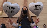 A woman in a T-shirt and wearing a face mask holds two heart-shaped balloons in her hands while posing for the camera. The one on her right reads "STOP THE HATE." The one on the left reads: "LOVE EACH OTHER."