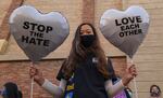 A woman in a T-shirt and wearing a face mask holds two heart-shaped balloons in her hands while posing for the camera. The one on her right reads "STOP THE HATE." The one on the left reads: "LOVE EACH OTHER."