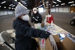 A woman in a hoodie, gloves and a winter hat reaches into a box of vaccine supplies while sitting at a table in a large, empty building. Another worker stands at the foot of the table while preparing doses.