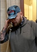 A man with a camouflaged hat embroidered with the letters "MAGA" speaks into a landline telephone.