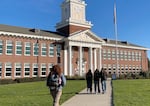 A blue sky and a brick building with a flag in the background. A group of three students and a single student behind them walk toward the school on the sidewalk.