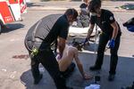 A person wearing no T-shirt sits in a parking lot as two people dressed in emergency responder clothes attend to his medical needs.