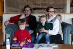 Malcolm and Simone Collins sit on a sofa in December 2024. Simone, seated on the right, has one child in her lap and another strapped to her back. Malcolm is holding one child, and their fourth child in on the floor in front of them, looking at a book open on a coffee table.