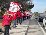 People dressed in winter gear, all wearing red, stand in a line outside a hospital. They're holding up signs that read "OFHNP Healthcare Workers On Strike Against Unfair Labor Practices," as cars drive past.