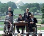 A man in a suit at a small desk leans over a piece of paper he signs. He is flanked on either side by four people. One is a man in a clerical collar. A woman with short hair wears a dark suit. Two men in suits are in wheelchairs. One wears a Western hat. They are outside on a green lawn. The white marble of the Thomas Jefferson Memorial is in the background.