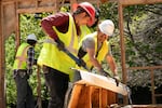 At a project site, two Perks Deconstruction crew members, wearing hard hats and yellow fluorescent vests, pull a white metal gutter from a piece of roofing that was taken off a house. The wooden frame of a structure stands in the background.