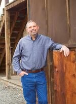 A man in blue jeans and a button down shirt leans against a wooden wall while smiling.