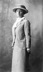 Black-and-white photo of a person in a skirt and jacket plus hat as she looks at the camera.