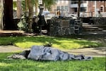 A person covered with a grey blanket sleeps on the grass of a park.