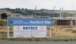 A sign reads "U.S. Department of Energy Hanford Site" and includes warnings that people and vehicles may be searched for prohibited items. In the background, a building and a crane can be seen.