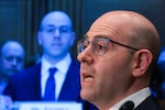 In this photo, Stephen Miran is seen from the neck up while testifying at a Senate Banking Committee hearing. He's wearing glasses and a shirt with a while collar.
