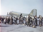 A group of men don jumpsuits in front of an airplane on a tarmac.