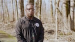 Darrell Wade stands with his held together in a wooded area. Portrait style.