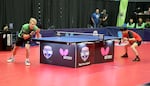 Two men play table tennis in a tournament in Portland. The table sits on a red floor.