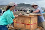 Two people drag a wooden stick across the top of a wooden box-like form, smoothing the sand inside. Spectators watch in the background.