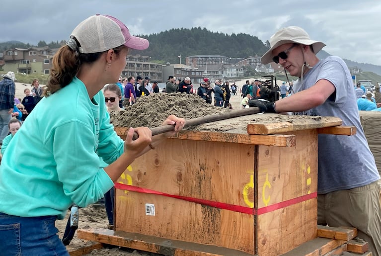 Two people drag a wooden stick across the top of a wooden box-like form, smoothing the sand inside. Spectators watch in the background.