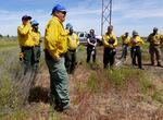 Man in a grassy field speaking to a circle of people, most of whom are wearing yellow fire shirts and green pants.