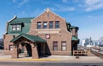 This photo shows Thief River Falls' City Hall, which used to be the Soo Line Railroad Depot. The two-story building is made of brown bricks and has a dark green metal roof. The right side of the frame shows a train track that leads to a grain elevator back in the distance.