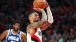 A man in Trail Blazers uniform lifts a basketball and prepares to shoot as a person behind him looks on wearing a 76ers uniform.