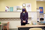 A woman wearing a mask and a business suit clasps her hands together while standing in an empty grade-school classroom.