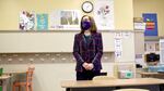 A woman wearing a mask and a business suit clasps her hands together while standing in an empty grade-school classroom.