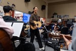 Standing with a guitar-like stringed instrument, Cindy Flores smiles as she teaches a mariachi class at McKay High School in Salem, Oregon. She is surrounded by seated students who are playing stringed instruments.
