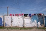 Pink and purple clothes hang on the top of a metal fence above tattered white and blue tents and tarps at Roj.