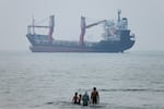 FILE — A group of people stands in shallow water as a cargo ship appears anchored in the Strait of Hormuz off Bandar Abbas, Iran, Tuesday, June 30, 2026.