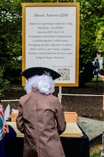This photo shows a sign outdoors that explains what America250 is. A person costumed in colonial attire, include a tricorne hat and white wig, is facing the sign.
