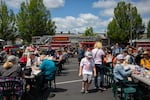 People sit at folding tables in a parking lot eating barbecue. A fire truck is parked on the street in the background.