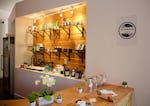 The interior of a clean and well-lit story shows a wood countertop and display area surrounded by white walls. On the countertop are flowers and other decorative plants and jars of candy. In the display area, boxed product are arrayed on shelves.