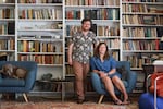 Ryan Holley is standing next to Annie Platt, who is seated in an armchair. Behind them are tall bookcases filled with books. At the left side of the frame, a dog is resting on an armchair.