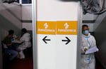 A small wall in the middle of the picture features two signs with the AstraZeneca logo and name on them. Arrows pointing in opposite directions are under each sign. To the right of the wall, a medical worker in a mask, protective eyewear and a white hazmat-style suit walks out of the room with papers in their hand. To the left of the wall, another white-suit clad worker sits at a table with a person in street clothes.